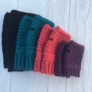 May include: Five pairs of crocheted fingerless gloves in different colors: black, teal, coral, purple, and a darker purple. The gloves are stacked on top of each other.