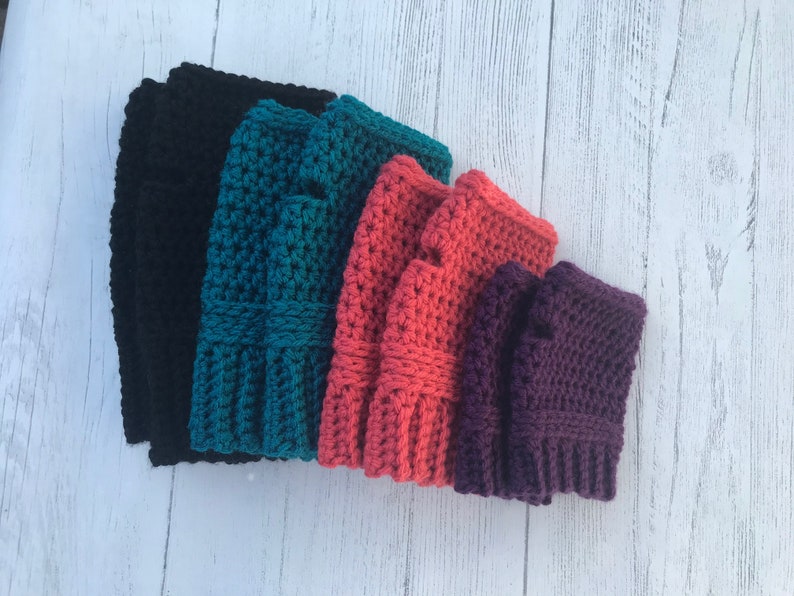 May include: Five pairs of crocheted fingerless gloves in different colors: black, teal, coral, purple, and a darker purple. The gloves are stacked on top of each other.