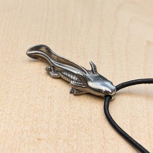 May include: A silver-colored axolotl pendant on a black cord necklace. The pendant is detailed with textured scales and fins. The necklace is displayed on a light brown wooden surface.