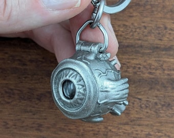 Puede incluir: Un llavero de metal plateado con un relicario esférico en forma de globo ocular. La superficie muestra detalles esculpidos intrincados, incluyendo un iris central, una pupila y venas texturizadas. El relicario está unido a una anilla metálica.