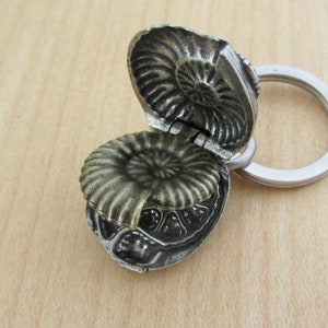 May include: A detailed, openwork metal keychain pendant shaped like an ammonite fossil. The pendant is a mix of silver and bronze tones, with a hinged top revealing the interior. The keychain ring is silver.