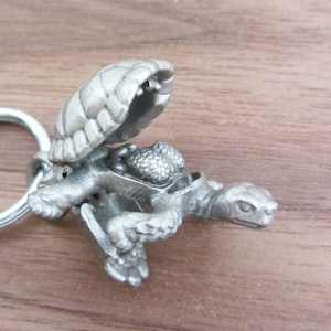 May include: A silver-colored turtle keychain with an open shell, revealing a textured interior. The turtle has detailed features and is attached to a silver key ring. The keychain is made of metal and is on a wooden surface.