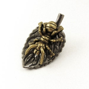 May include: A detailed metal brooch featuring a spider resting on a leaf. The spider has a gold-colored body and legs, while the leaf is a darker, silver-toned metal. The brooch is designed with intricate details.