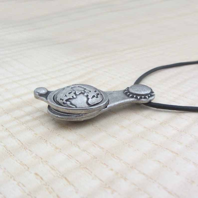 May include: A silver pendant necklace with a globe design. The pendant is attached to a black cord.