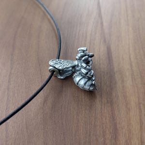 May include: A silver bee pendant necklace with a black leather cord. The bee is detailed with wings, legs, and a stinger.