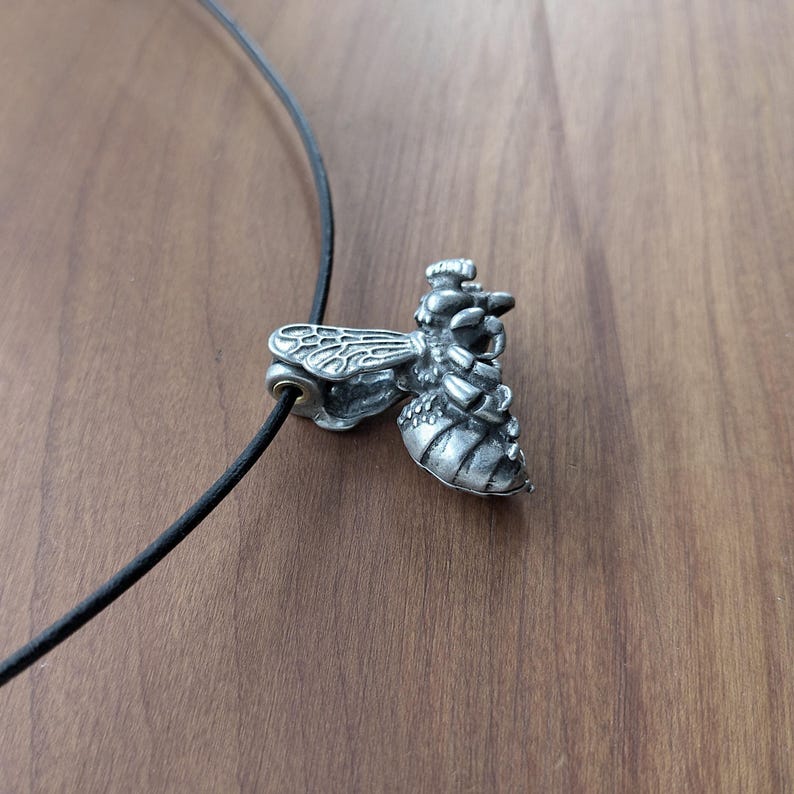 May include: A silver bee pendant necklace with a black leather cord. The bee is detailed with wings, legs, and a stinger.