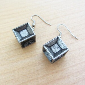 May include: A pair of silver-colored cube earrings with a geometric design. Each earring features a square shape with a recessed diamond pattern in the center. The earrings hang from silver-colored fish hook ear wires.