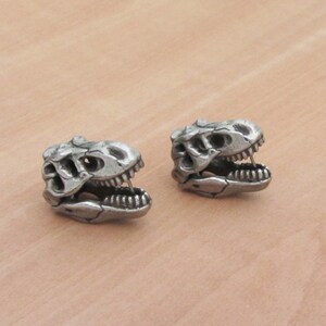 May include: Two silver-colored dinosaur skull-shaped stud earrings. The earrings are detailed with visible teeth and eye sockets, and are set against a neutral background. The earrings are likely made of metal.