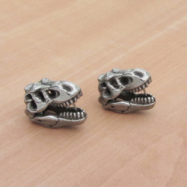 May include: Two silver-colored dinosaur skull-shaped stud earrings. The earrings are detailed with visible teeth and eye sockets, and are set against a neutral background. The earrings are likely made of metal.