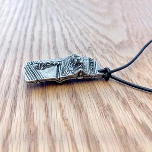 Op de afbeelding: Een zilverkleurige hanger met ingewikkelde reli&euml;fdetails, waaronder verticale lijnen en sierlijke patronen. De rechthoekige hanger heeft een gedetailleerde bovenrand en is bevestigd aan een zwarte koordketting. De hanger wordt op een houten oppervlak gepresenteerd.