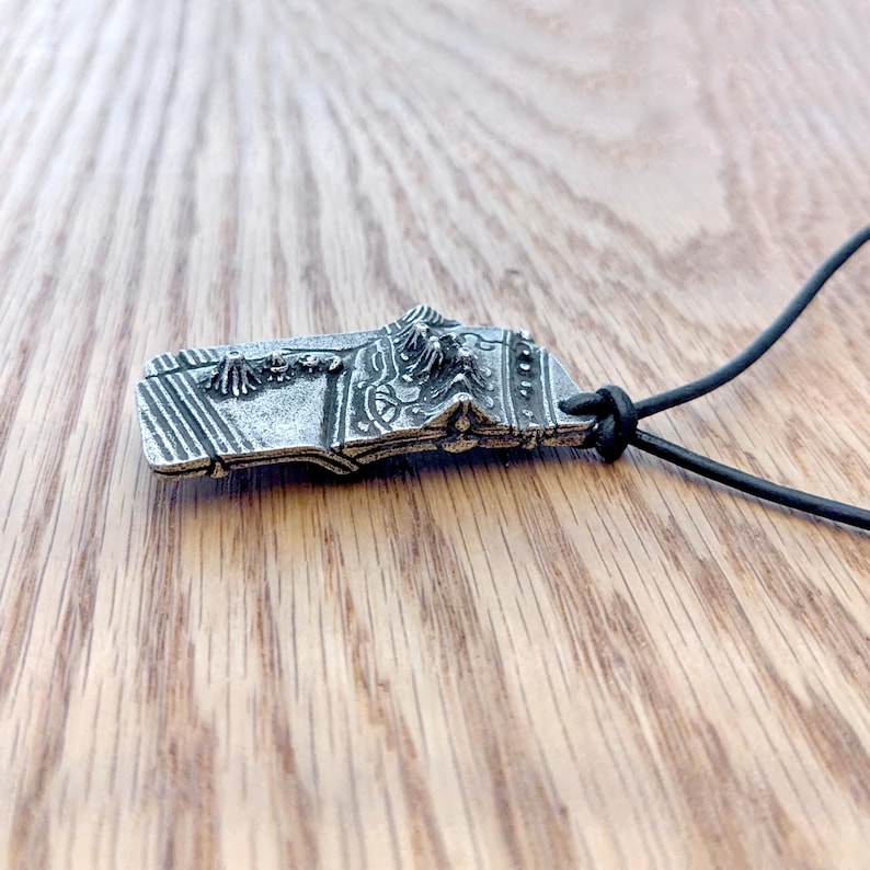 Op de afbeelding: Een zilverkleurige hanger met ingewikkelde reli&euml;fdetails, waaronder verticale lijnen en sierlijke patronen. De rechthoekige hanger heeft een gedetailleerde bovenrand en is bevestigd aan een zwarte koordketting. De hanger wordt op een houten oppervlak gepresenteerd.