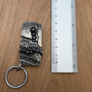 May include: A silver-colored rectangular keychain with an ornate, textured design. The keychain is next to a ruler, showing its length. The design includes floral and geometric patterns. The keychain's length is approximately 2 inches.