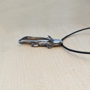May include: A silver-colored axolotl pendant on a black cord. The pendant is detailed with visible gills and a textured body. The axolotl is a salamander known for its unique appearance. The cord is thin and appears to be made of a rubber-like material.