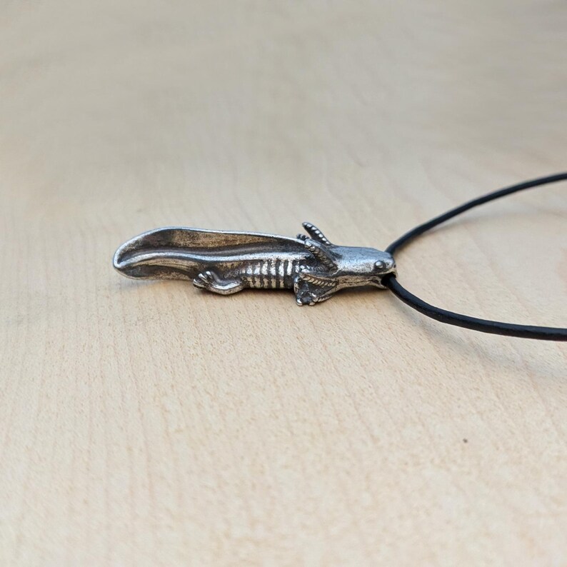 May include: A silver-colored axolotl pendant on a black cord. The pendant is detailed with visible gills and a textured body. The axolotl is a salamander known for its unique appearance. The cord is thin and appears to be made of a rubber-like material.