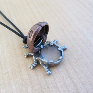 May include: A unique pendant necklace featuring a detailed, open-mouthed bug design. The pendant has a copper-colored head and a silver-colored body with a circular, light-colored stone. The necklace has a black cord.
