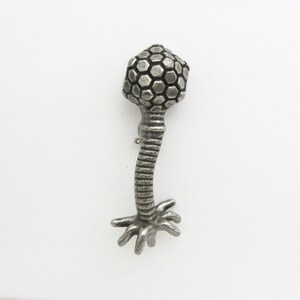 Może przedstawiać: Srebrna metalowa broszka w kształcie wirusa bakteriofaga. Broszka ma sześciokątną głowę, żebrowany trzon i podstawę z wieloma mackami. Projekt jest szczeg&oacute;łowy i tr&oacute;jwymiarowy.