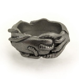 May include: A dark gray ring with a textured surface. The ring features a design of two hands clasped together, creating a unique and artistic look. The ring is made of a solid material and has a matte finish.