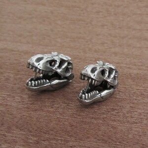 May include: Two silver-colored dinosaur skull figurines. The skulls have detailed features, including teeth and eye sockets. The figurines are displayed on a brown wooden surface.