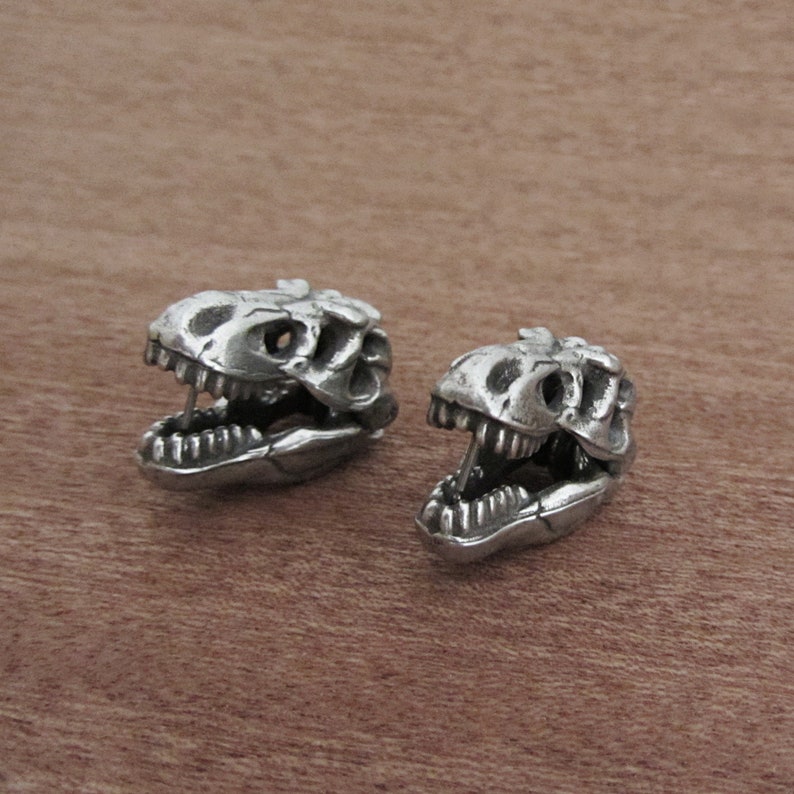 May include: Two silver-colored dinosaur skull figurines. The skulls have detailed features, including teeth and eye sockets. The figurines are displayed on a brown wooden surface.