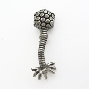 Może przedstawiać: Srebrny metalowy wisiorek w kształcie wirusa bakteriofaga. Wisiorek ma sześciokątną głowę, żebrowaną szyję i podstawę z wieloma mackami. Projekt jest szczeg&oacute;łowy i tr&oacute;jwymiarowy, z lekko postarzanym wyglądem.