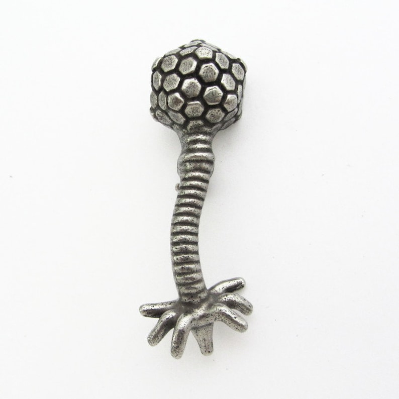 Może przedstawiać: Srebrny metalowy wisiorek w kształcie wirusa bakteriofaga. Wisiorek ma sześciokątną głowę, żebrowaną szyję i podstawę z wieloma mackami. Projekt jest szczeg&oacute;łowy i tr&oacute;jwymiarowy, z lekko postarzanym wyglądem.