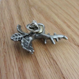 May include: A silver-toned metal lapel pin in the shape of a stylized plant with leaves and a branch-like structure. The pin has a textured surface and a pin back for attachment. The pin is set against a wooden background.