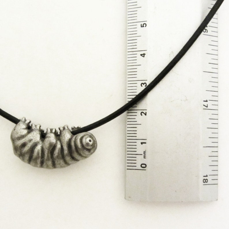 May include: A silver-toned pendant necklace featuring a detailed, stylized caterpillar charm. The caterpillar is suspended from a black cord. A ruler is placed next to the pendant for scale. The pendant measures approximately 1 inch in length.