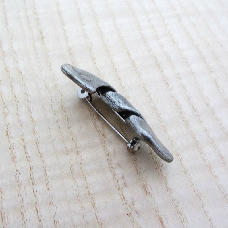 May include: A silver-toned metal brooch with a textured, segmented design. The brooch has a pin closure on the back. The design is elongated and slightly curved, with a matte finish. The brooch is likely a fashion accessory.