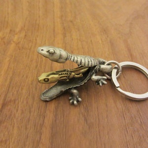 May include: A metal keychain with a lizard and snake design. The lizard is silver, and the snake is gold. The keychain has a silver key ring.