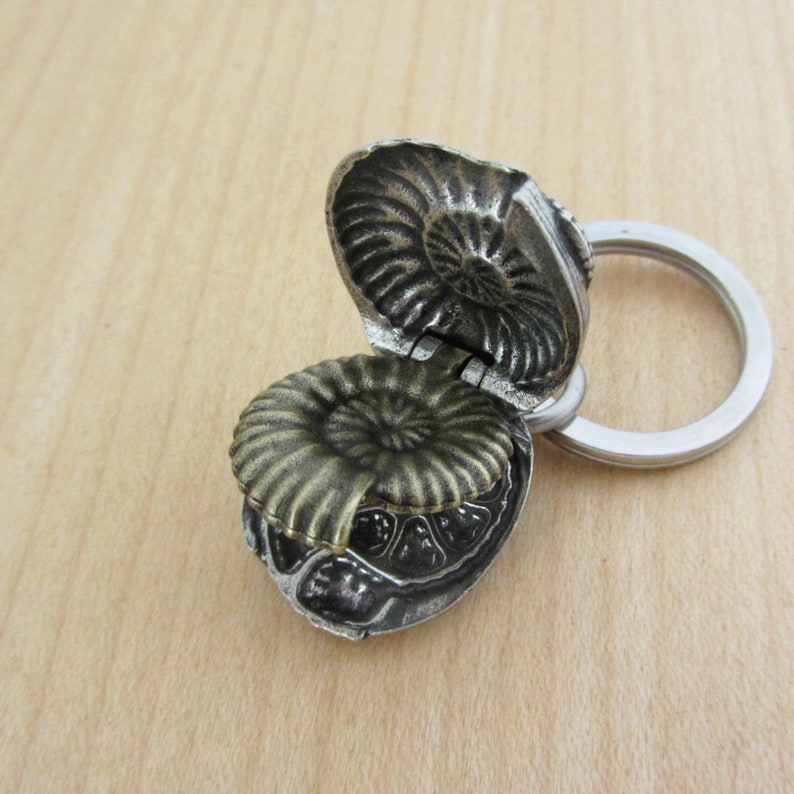 May include: A detailed, openwork metal keychain pendant shaped like an ammonite fossil. The pendant is a mix of silver and bronze tones, with a hinged top revealing the interior. The keychain ring is silver.