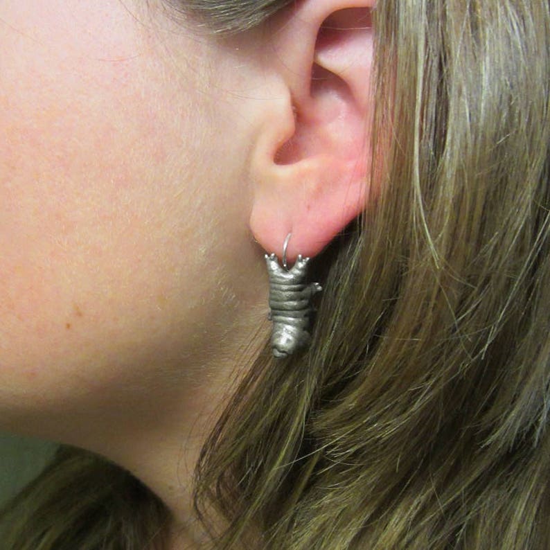 May include: A silver-toned metal earring shaped like a tardigrade, also known as a water bear. The creature has a segmented body, eight small legs, and a rounded head, hanging from a simple silver-colored metal hoop wire in the earlobe.