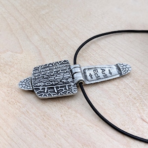 Puede incluir: Collar con colgante plateado y cord&oacute;n negro. El colgante presenta un dise&ntilde;o rectangular con un patr&oacute;n texturizado y el texto "ATM Ecm". El colgante est&aacute; conectado al cord&oacute;n con un mecanismo de bisagra. Una joya elegante.