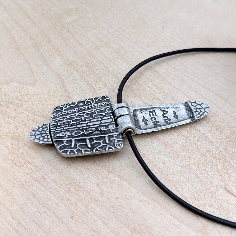 Puede incluir: Collar con colgante plateado y cord&oacute;n negro. El colgante presenta un dise&ntilde;o rectangular con un patr&oacute;n texturizado y el texto "ATM Ecm". El colgante est&aacute; conectado al cord&oacute;n con un mecanismo de bisagra. Una joya elegante.