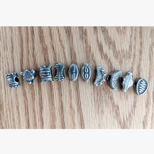 May include: A collection of ten individual silver-toned metal beads arranged in a horizontal row. Each bead features a unique embossed pattern, including geometric lines, textured surfaces, and varied shapes such as cylinders and crescents.