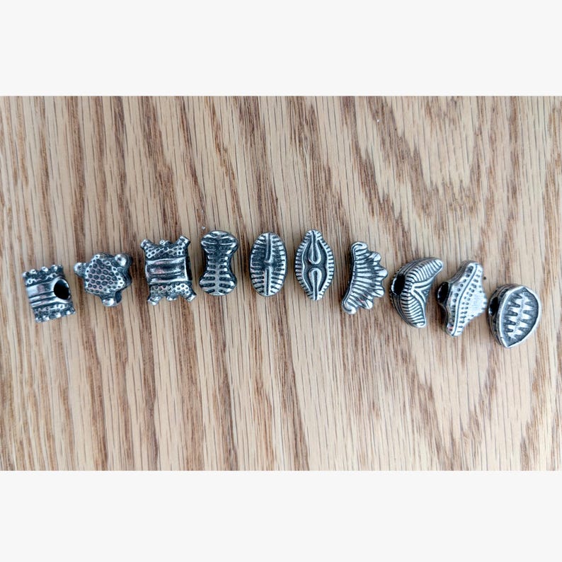 May include: A collection of ten individual silver-toned metal beads arranged in a horizontal row. Each bead features a unique embossed pattern, including geometric lines, textured surfaces, and varied shapes such as cylinders and crescents.