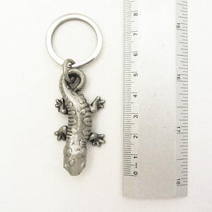 May include: A silver-coloured metal keyring featuring a detailed lizard pendant. The lizard has a textured body and four small feet. The keyring includes a metal ring for attaching keys. The lizard pendant measures approximately 5 cm in length.