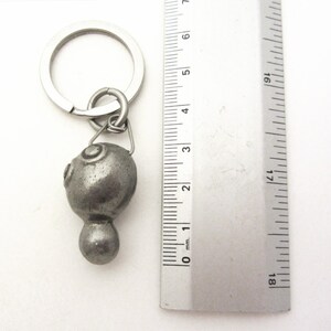 May include: A silver-colored metal keychain with a decorative, rounded pendant. The pendant has a textured surface with two circular designs and a small, rounded base. The keychain includes a metal ring and a connecting loop. The ruler next to the keychain shows the pendant is approximately 1 inch in length.