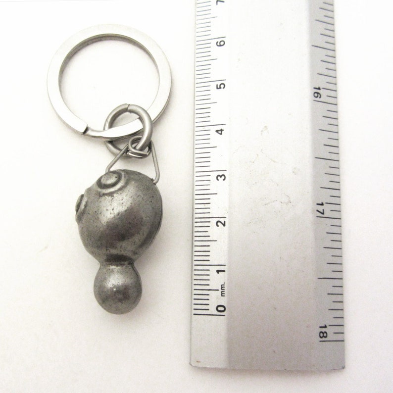 May include: A silver-colored metal keychain with a decorative, rounded pendant. The pendant has a textured surface with two circular designs and a small, rounded base. The keychain includes a metal ring and a connecting loop. The ruler next to the keychain shows the pendant is approximately 1 inch in length.