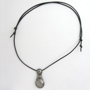 May include: A silver pendant necklace with a round, detailed design hangs from a black leather cord.