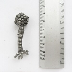 May include: A silver-toned metal pin shaped like a bacteriophage, a type of virus that infects bacteria. The pin has a round head with a hexagonal pattern and a long, thin tail with a base that resembles a set of roots.