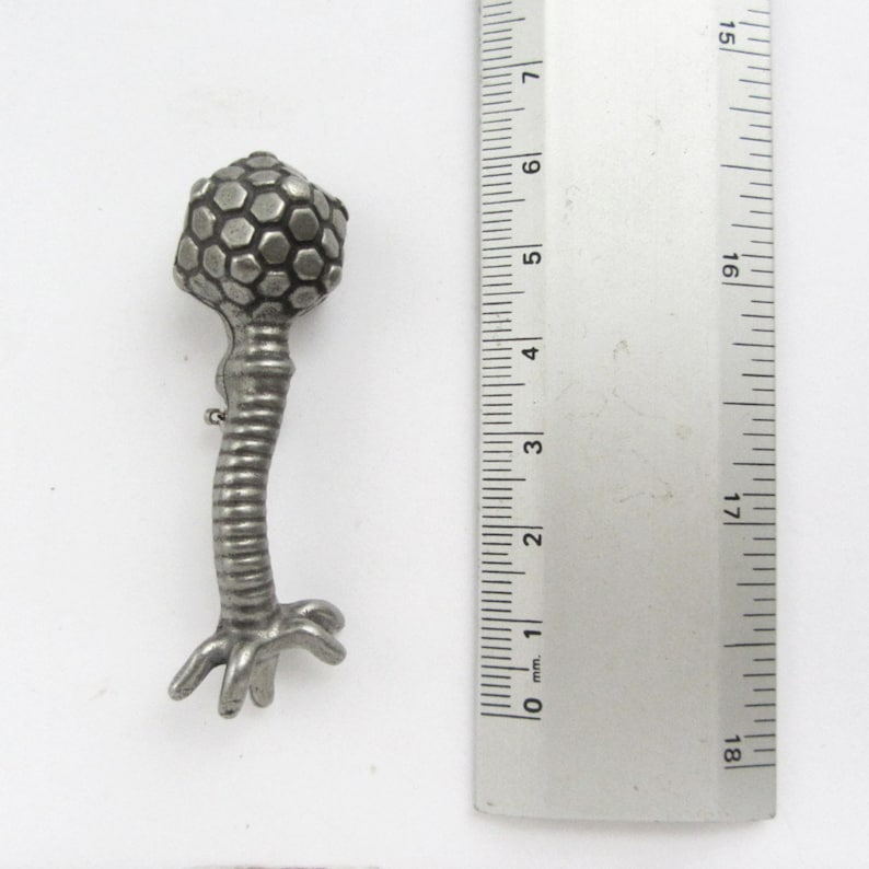 May include: A silver-toned metal pin shaped like a bacteriophage, a type of virus that infects bacteria. The pin has a round head with a hexagonal pattern and a long, thin tail with a base that resembles a set of roots.