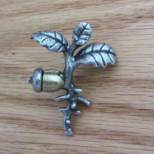 May include: A silver-toned metal acorn and sprout decorative object. The acorn is gold-colored, and the sprout has three leaves. The object is set against a wooden background.