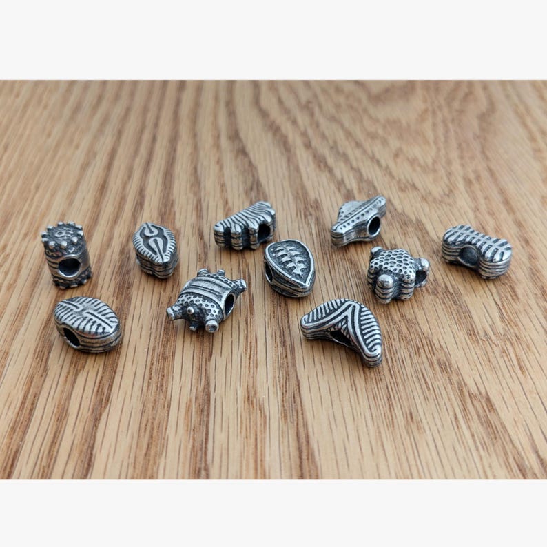 May include: A collection of eleven small, silver-toned metal beads featuring various embossed geometric patterns, including stripes, dots, and ridges. Each bead has a central hole for threading, with an oxidized finish that highlights the intricate textures.