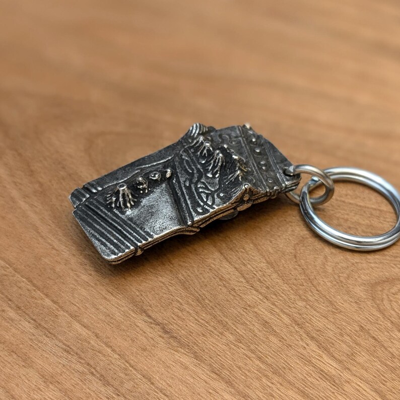 May include: A silver-colored rectangular keychain with a detailed relief design. The design appears to depict a castle or architectural structure. The keychain has a large, round key ring attached.