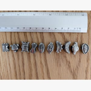 May include: Ten silver-toned metal beads with various embossed patterns and shapes arranged in a horizontal row. A white plastic ruler is positioned above the beads, showing that each individual bead measures approximately 0.5 inches in width.