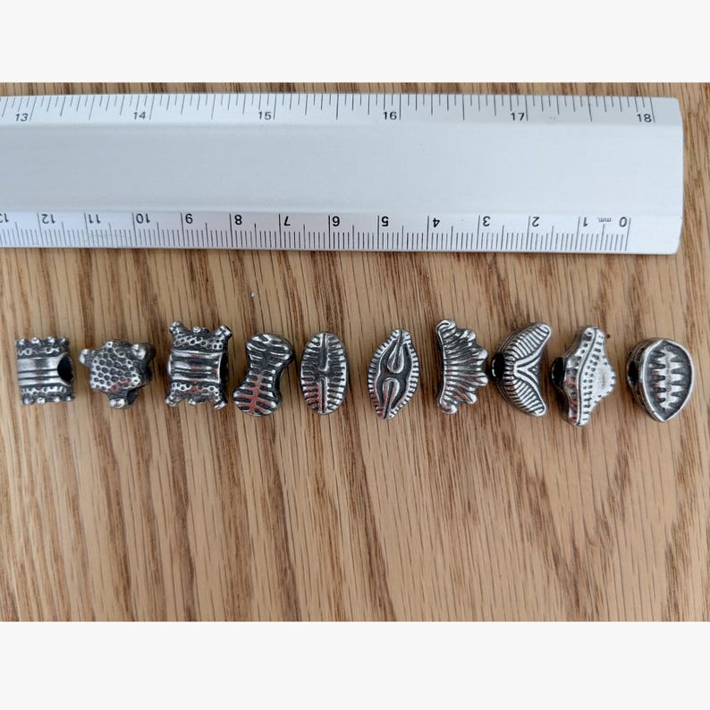 May include: Ten silver-toned metal beads with various embossed patterns and shapes arranged in a horizontal row. A white plastic ruler is positioned above the beads, showing that each individual bead measures approximately 0.5 inches in width.