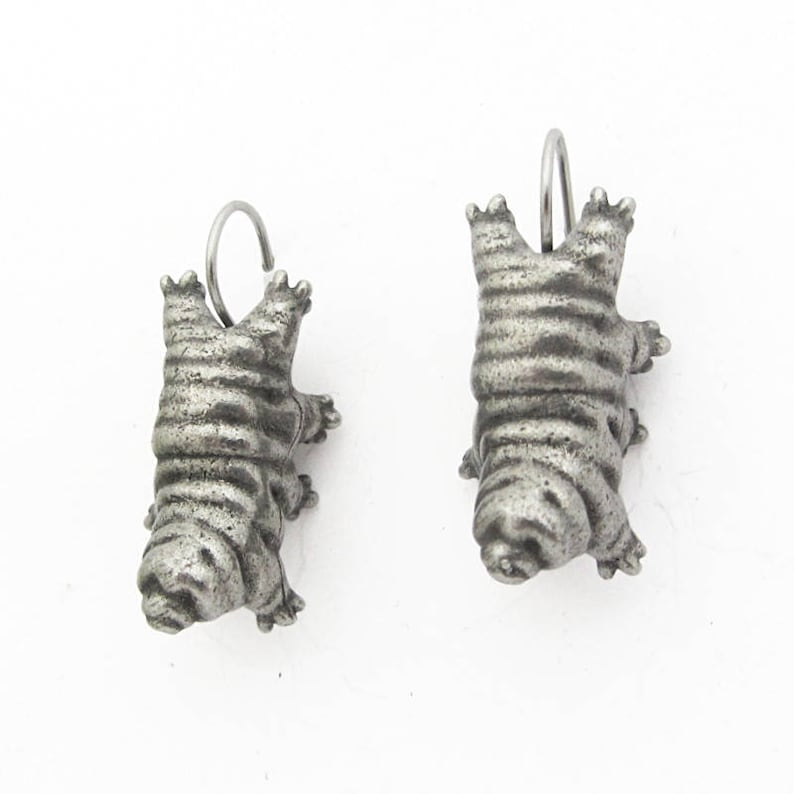 May include: A pair of silver-toned metal earrings shaped like tardigrades, also known as water bears. Each earring features a segmented, plump body with eight small legs and a curved wire hook for attachment, showcasing a weathered, textured finish.