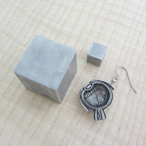 May include: A silver eye-shaped earring with a detailed design and a silver hook. The earring is displayed on a wooden surface with two small silver cubes.