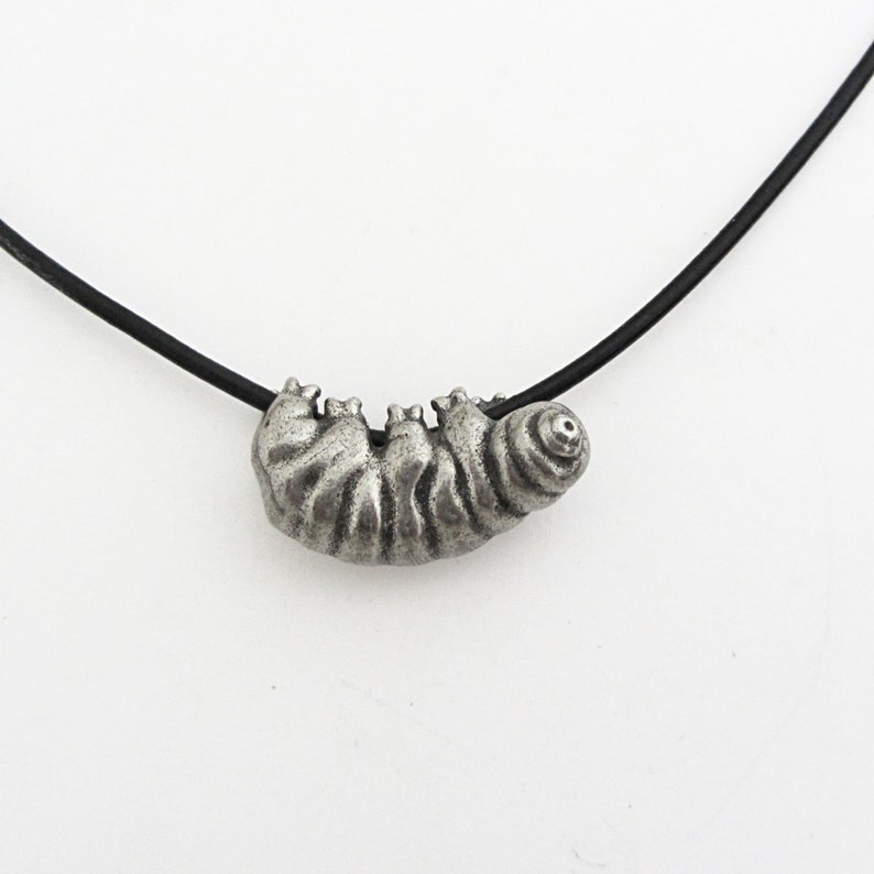 May include: A silver-colored caterpillar pendant necklace. The pendant is detailed with segmented body sections and sits on a black cord. The necklace is a unique piece of jewelry.