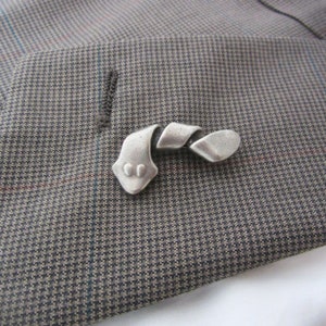 May include: A silver-toned metal lapel pin in a stylized, abstract design resembling a snake or ribbon. The pin is placed on a gray and brown patterned fabric, likely a suit jacket.